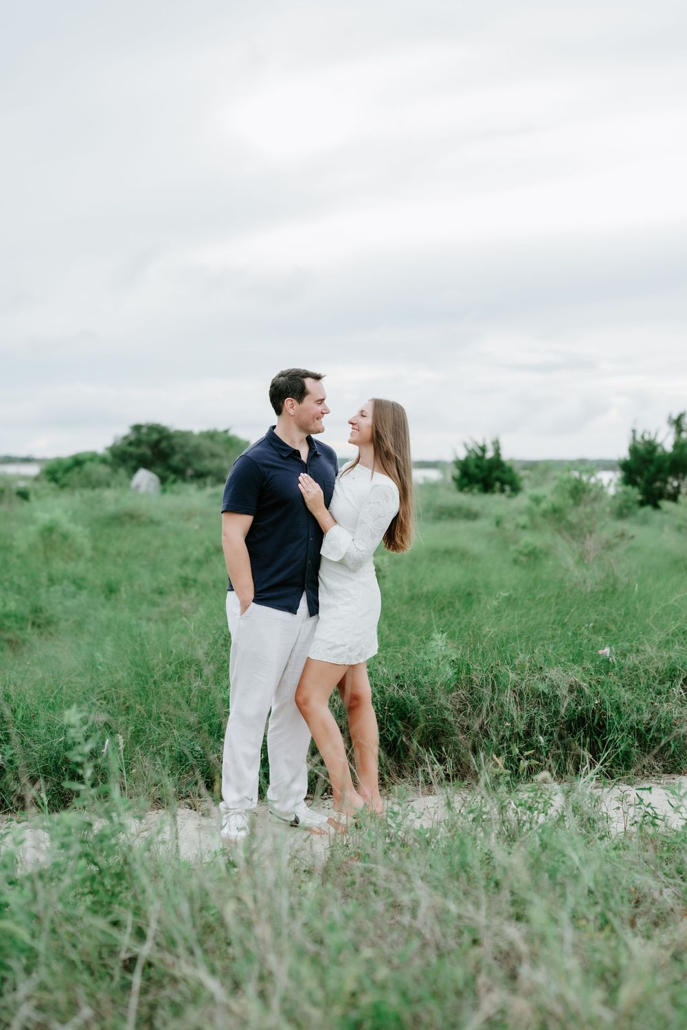 Couple embracing in a green field, woman in white dress and man in navy polo, sharing a romantic moment outdoors.