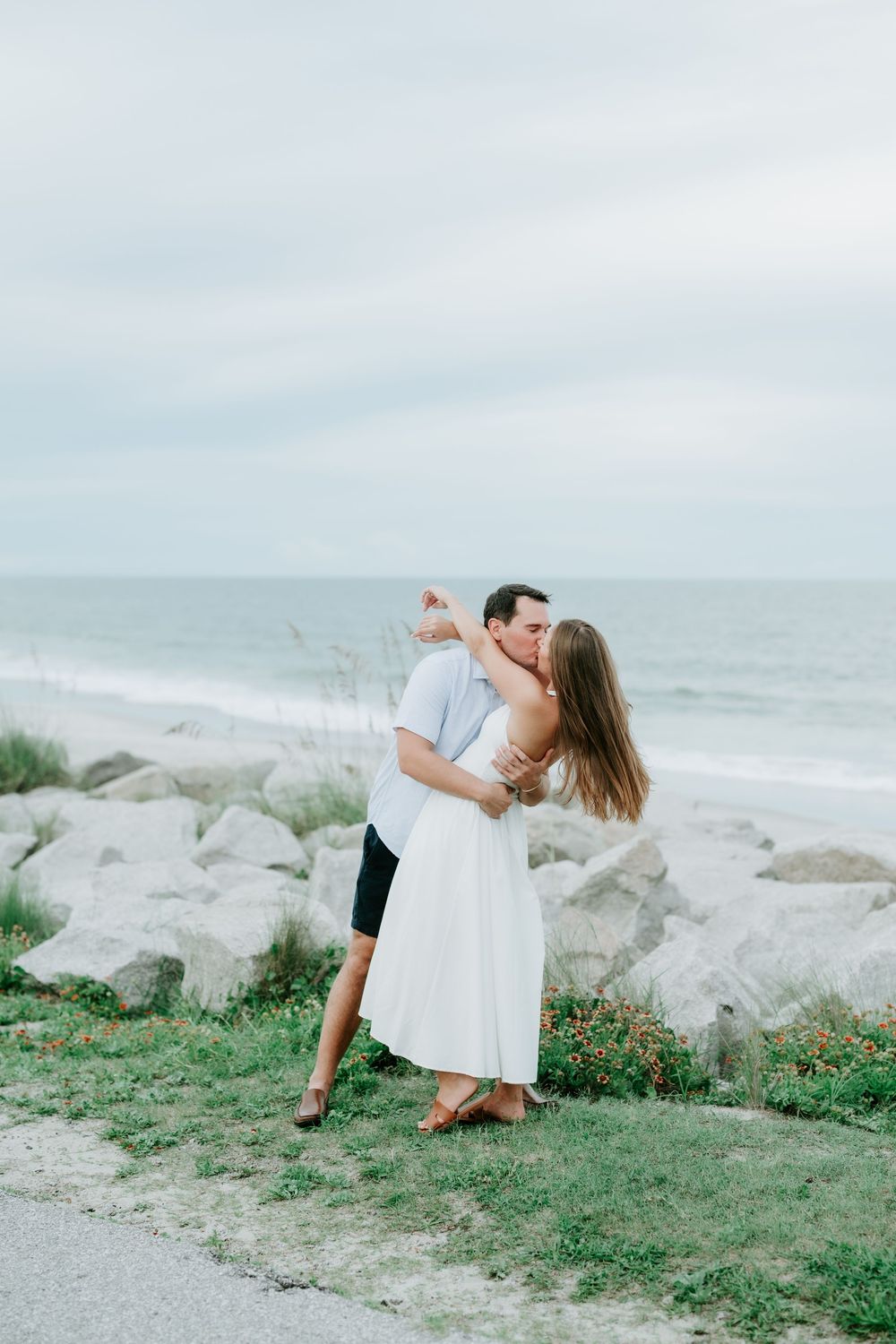 Couple sharing a romantic kiss near rocky shoreline with ocean backdrop, woman in white dress and man in blue shirt.