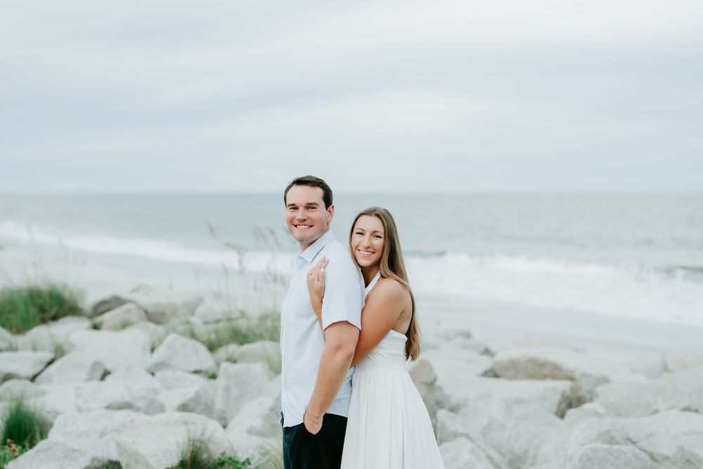 Couple in white outfits posing on rocky beach with ocean waves in background.