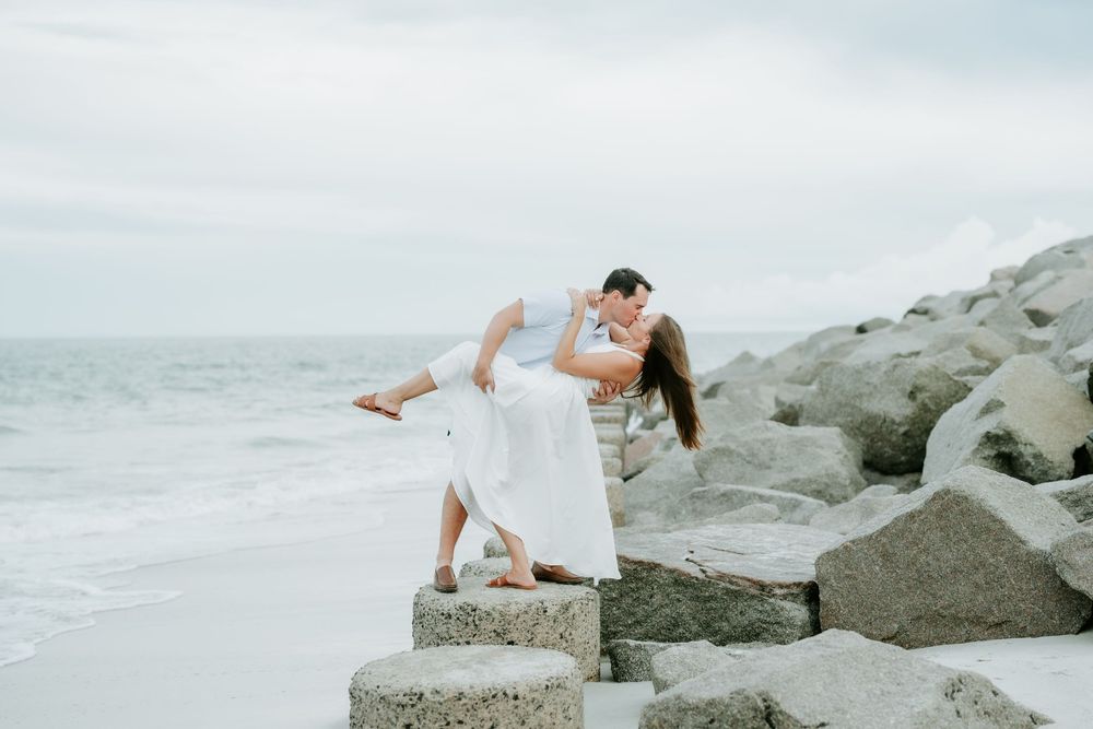 Couple sharing a romantic dip kiss on rocky beach with ocean waves in background, wearing white outfits.
