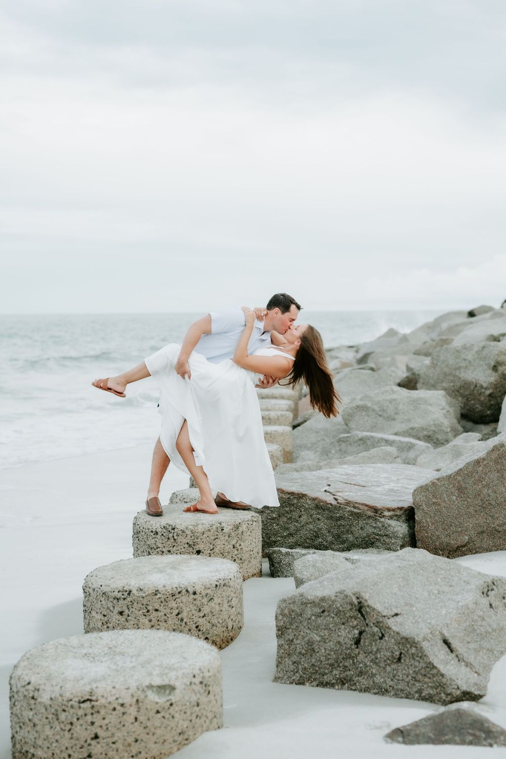 Couple sharing a romantic dip kiss on rocky jetty steps with ocean waves in the background.