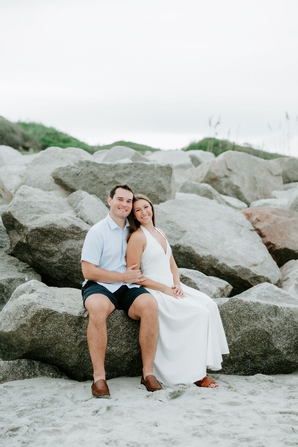 Happy couple sitting on large beach rocks, woman in white dress leaning on mans shoulder, smiling at camera.