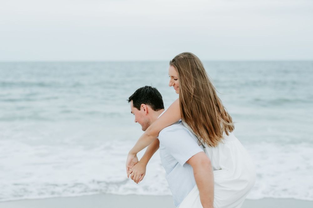 A couple shares a romantic piggyback ride on a beach, with ocean waves in the background.