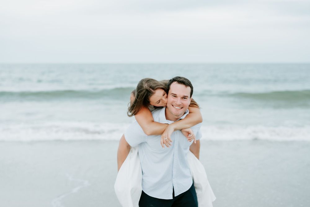 A smiling man gives a young girl a piggyback ride on a beach with ocean waves in the background.