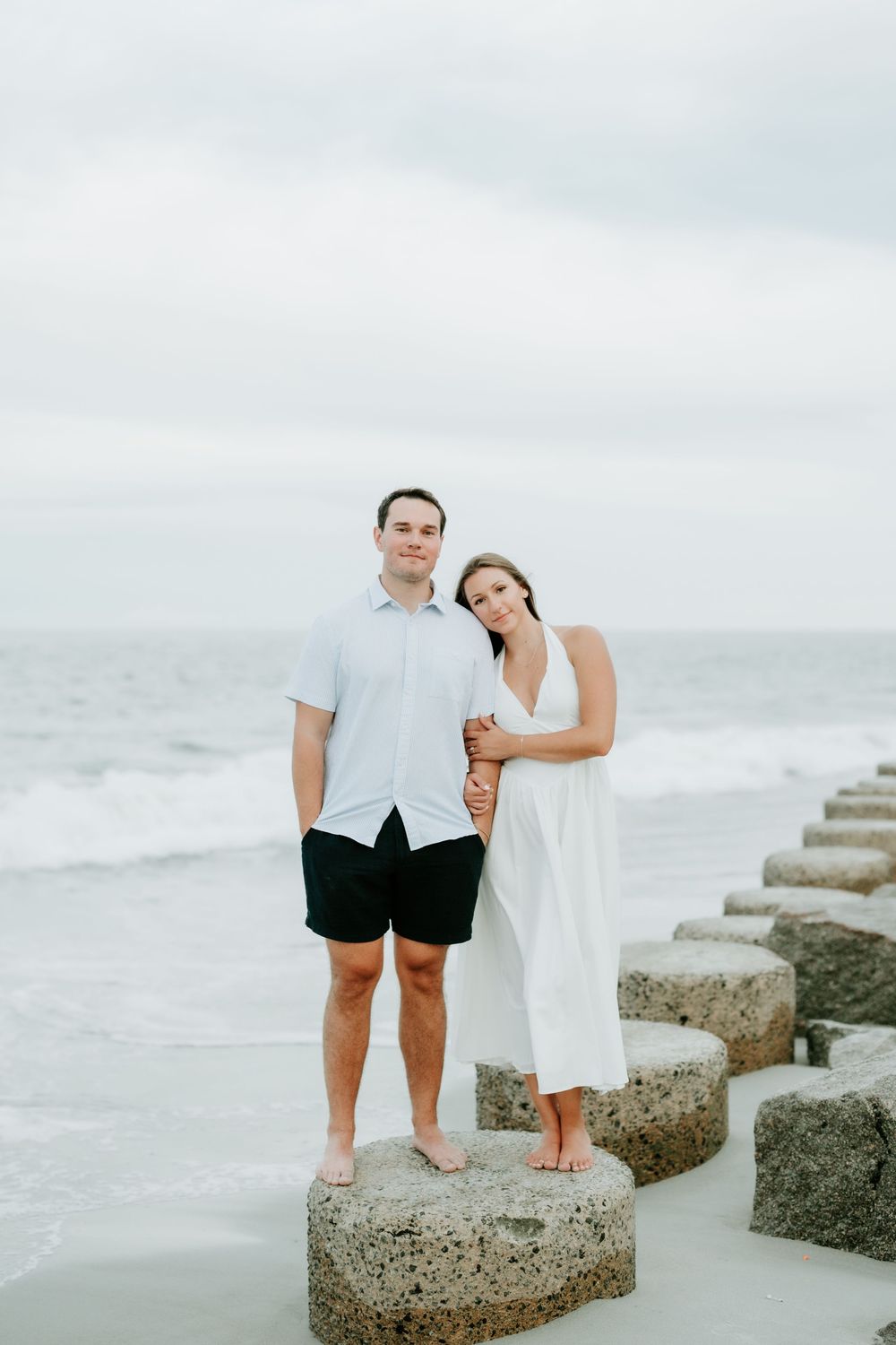 Couple posing on seaside rocks, woman in white dress leaning on man in white shirt and black shorts, ocean backdrop.