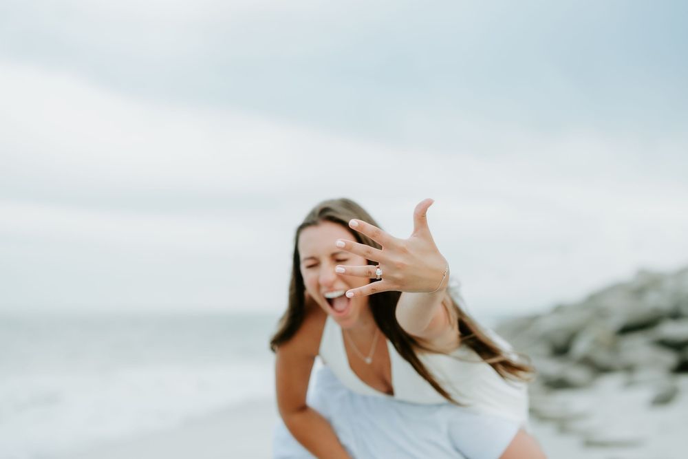 Woman laughing on beach showing off engagement ring while wearing white dress with ocean backdrop.