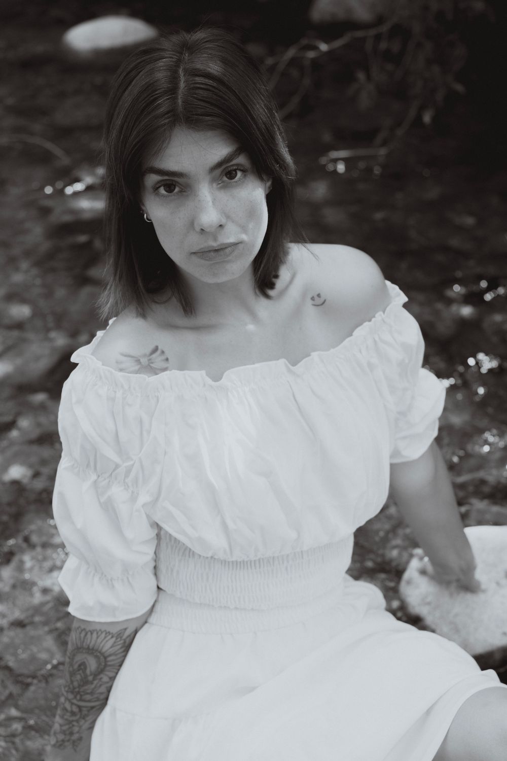 Black and white portrait of a young woman with tattoos wearing an off-shoulder white dress, sitting outdoors.