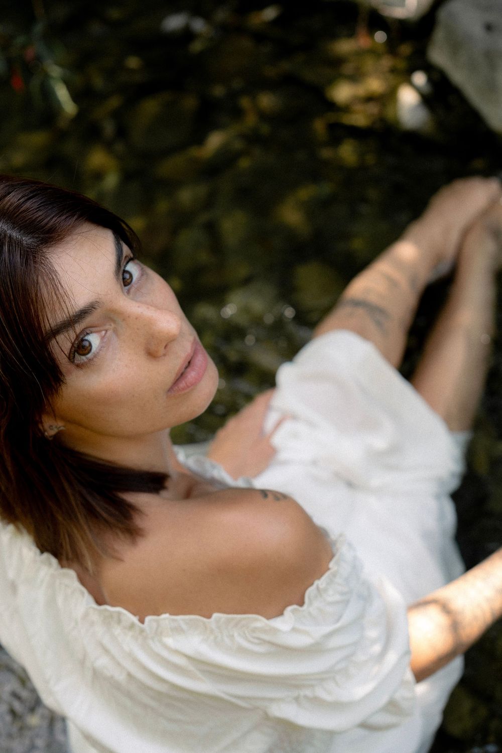 Woman in white off-shoulder dress with tattoos sits near water, gazing up with natural bokeh background.