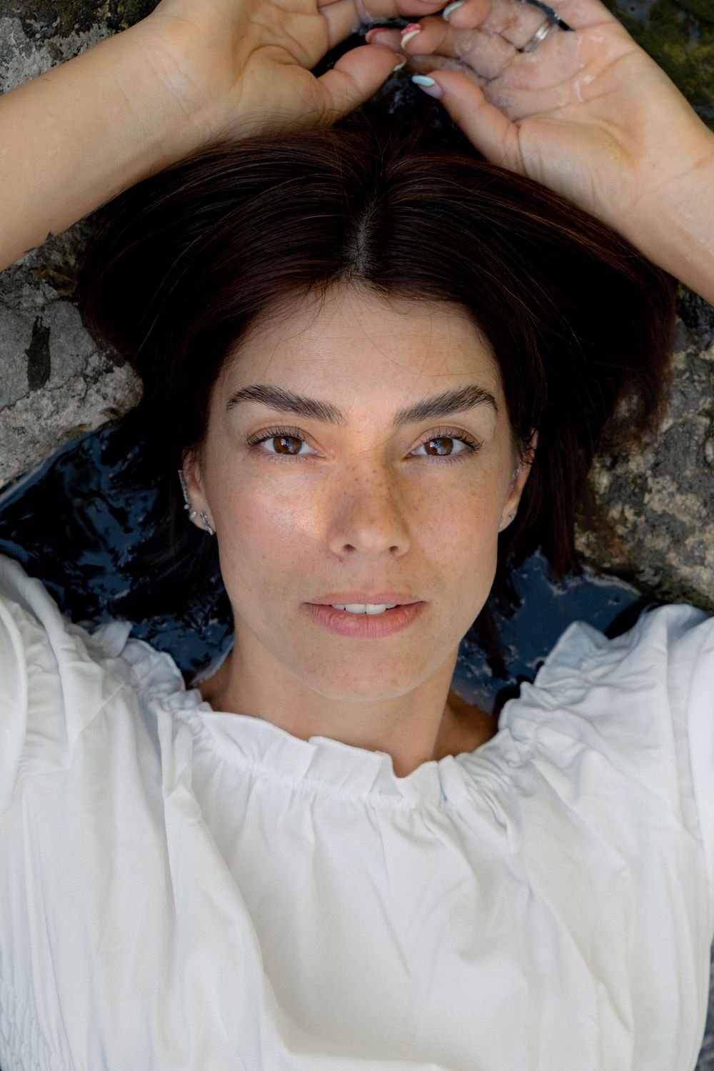 Woman with dark hair lying back, hands raised above head, wearing white blouse, looking at camera.