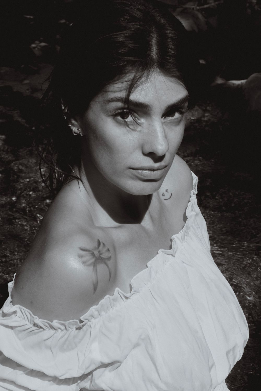 Black and white portrait of a young woman in an off-shoulder white dress with a floral tattoo, gazing upward.