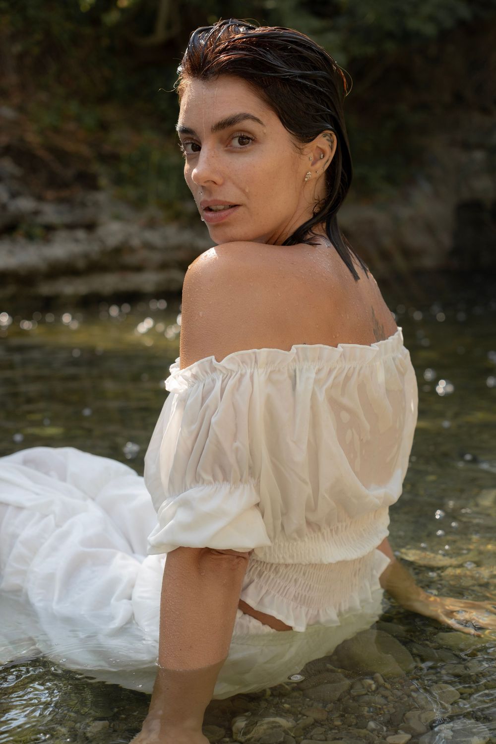 Woman in white off-shoulder dress sitting in a shallow stream, with wet hair and a serene expression.