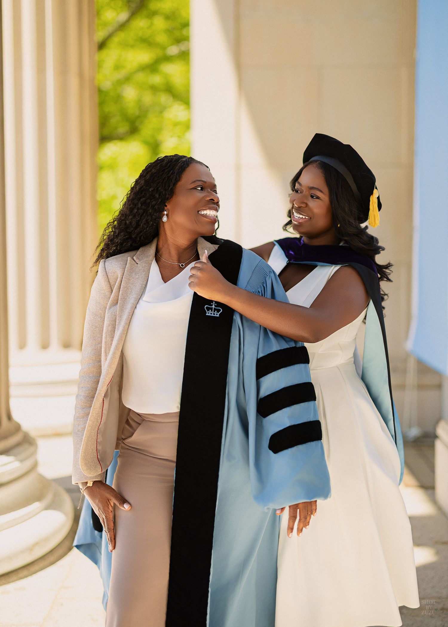 Two women in graduation regalia smile joyfully together near white columns, celebrating an academic achievement outdoors.