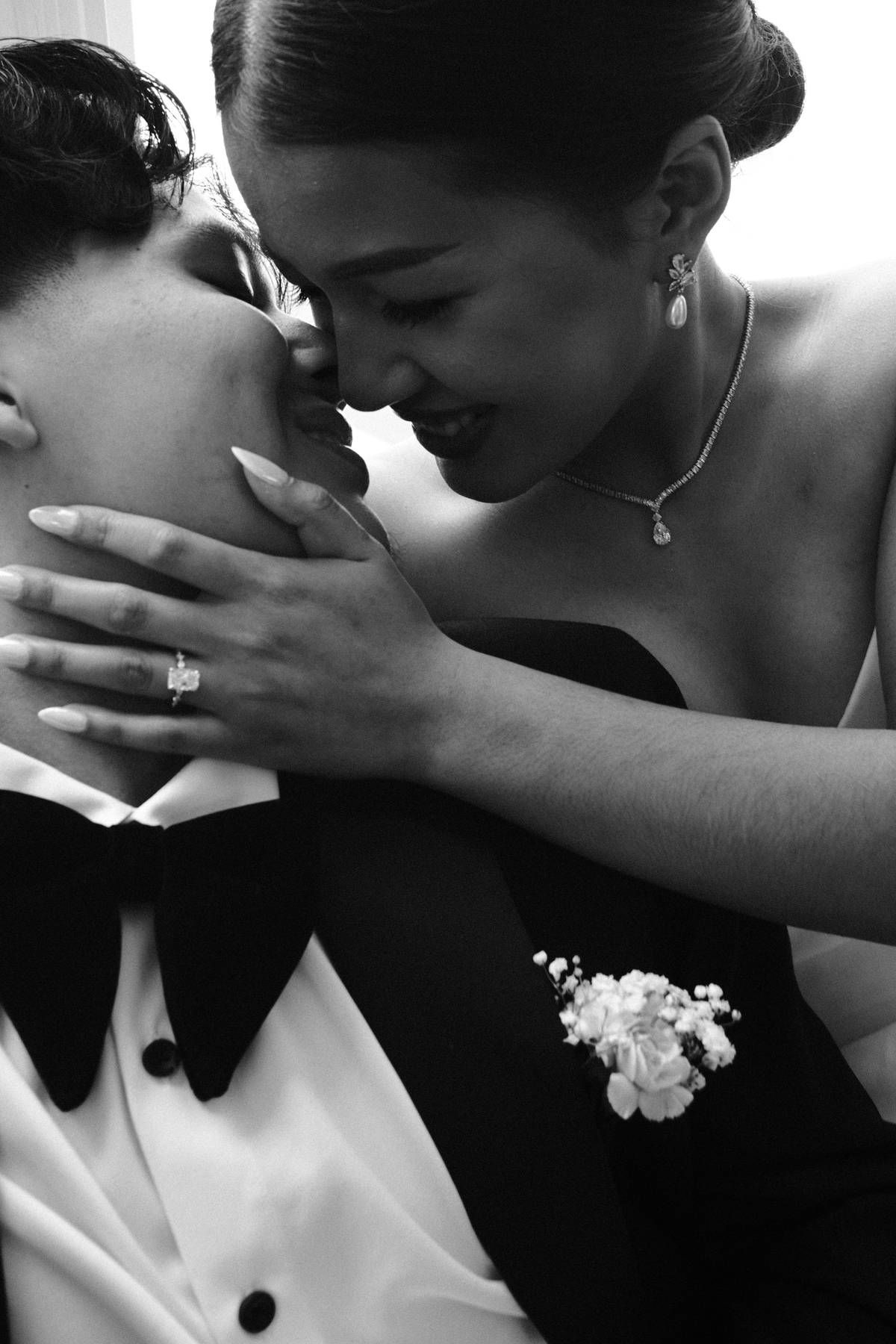 Black and white photo of a smiling bride leaning in to kiss groom in tuxedo with boutonniere on wedding day.