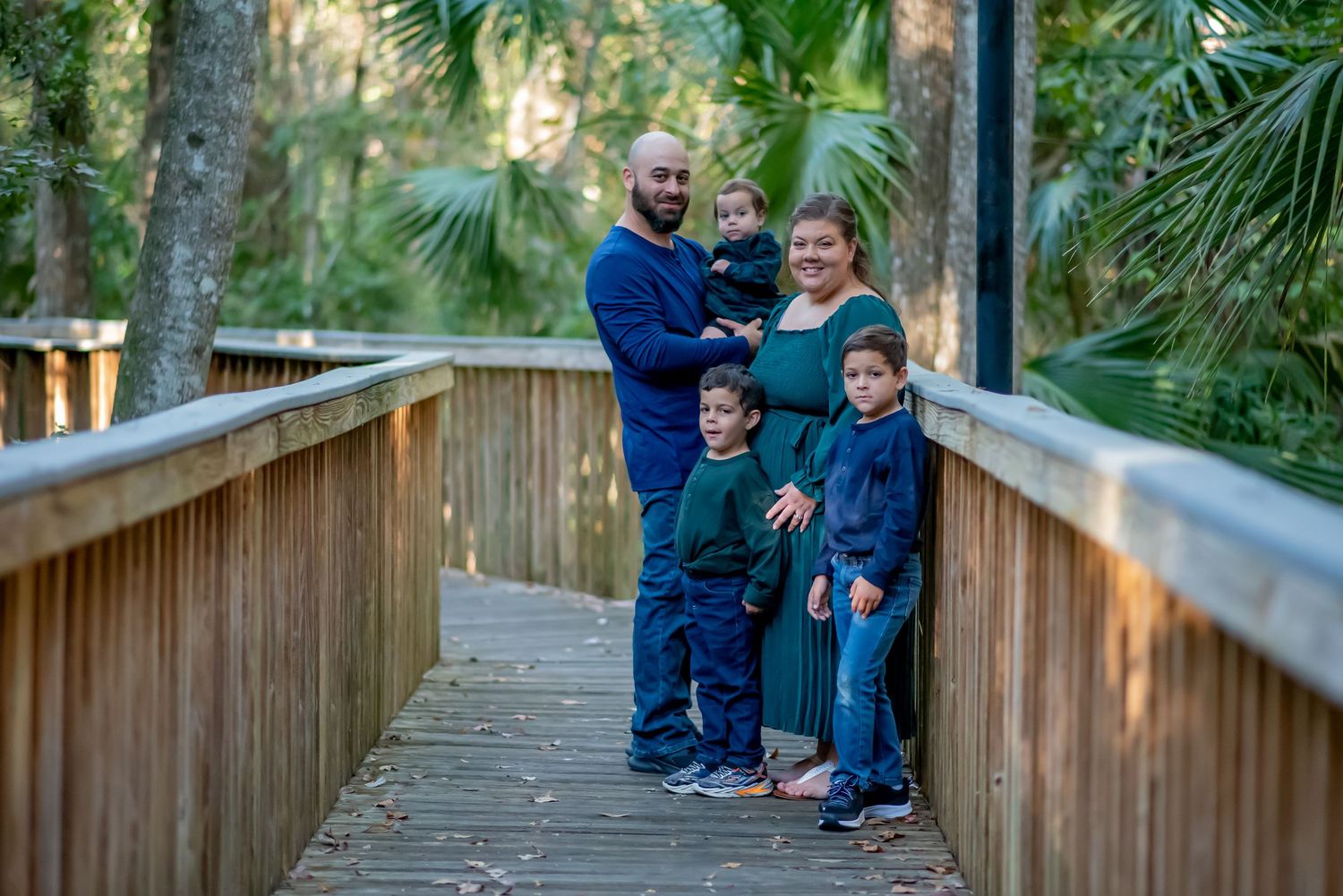 Family of five posing on a wooden boardwalk surrounded by lush green trees in a natural park setting.