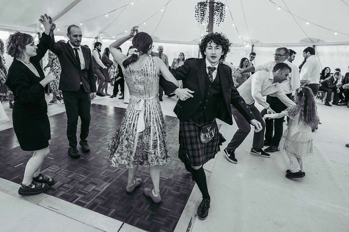 Black and white photo of guests dancing energetically at a wedding reception in a marquee tent.
