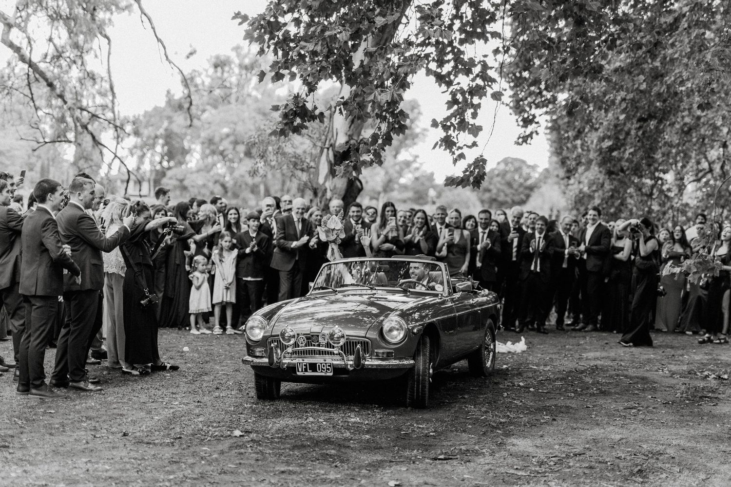 Black and white photo of newlyweds departing in a classic convertible car, surrounded by cheering wedding guests outdoors.
