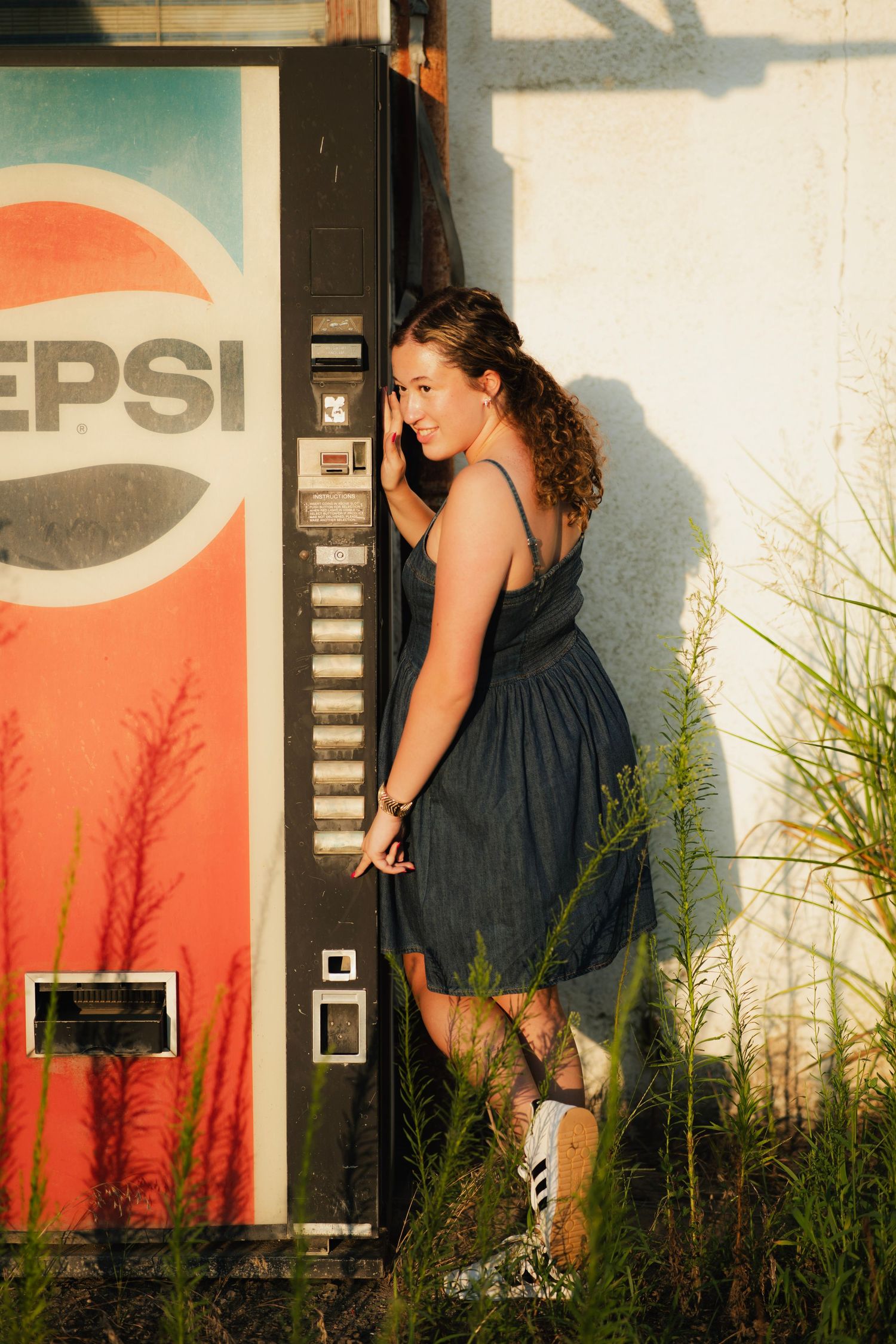 Young woman in dark dress posing beside a vintage Pepsi vending machine outdoors at golden hour.