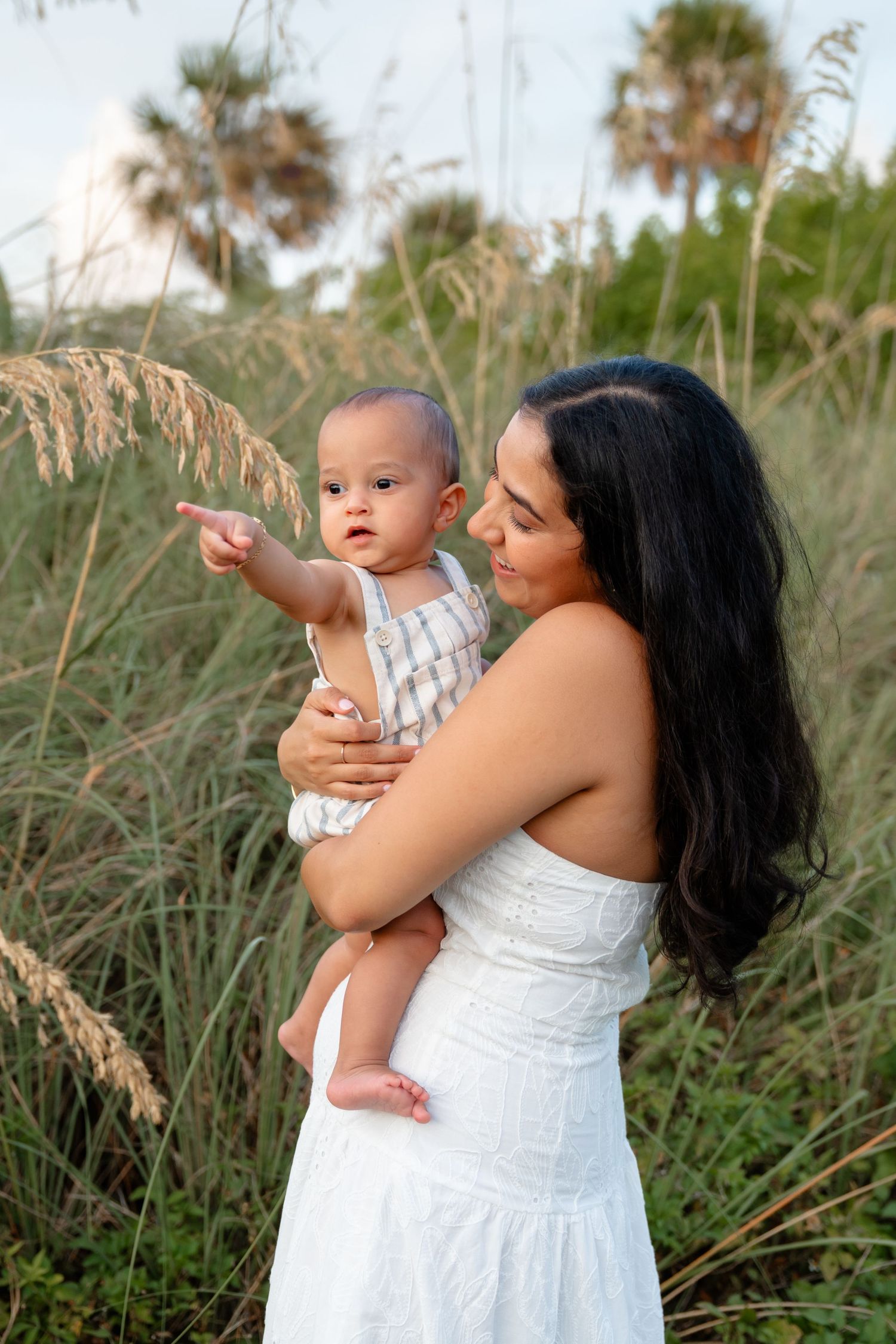 A mother in a white dress holds her baby outdoors among tall grass and palm trees, as the infant points curiously.