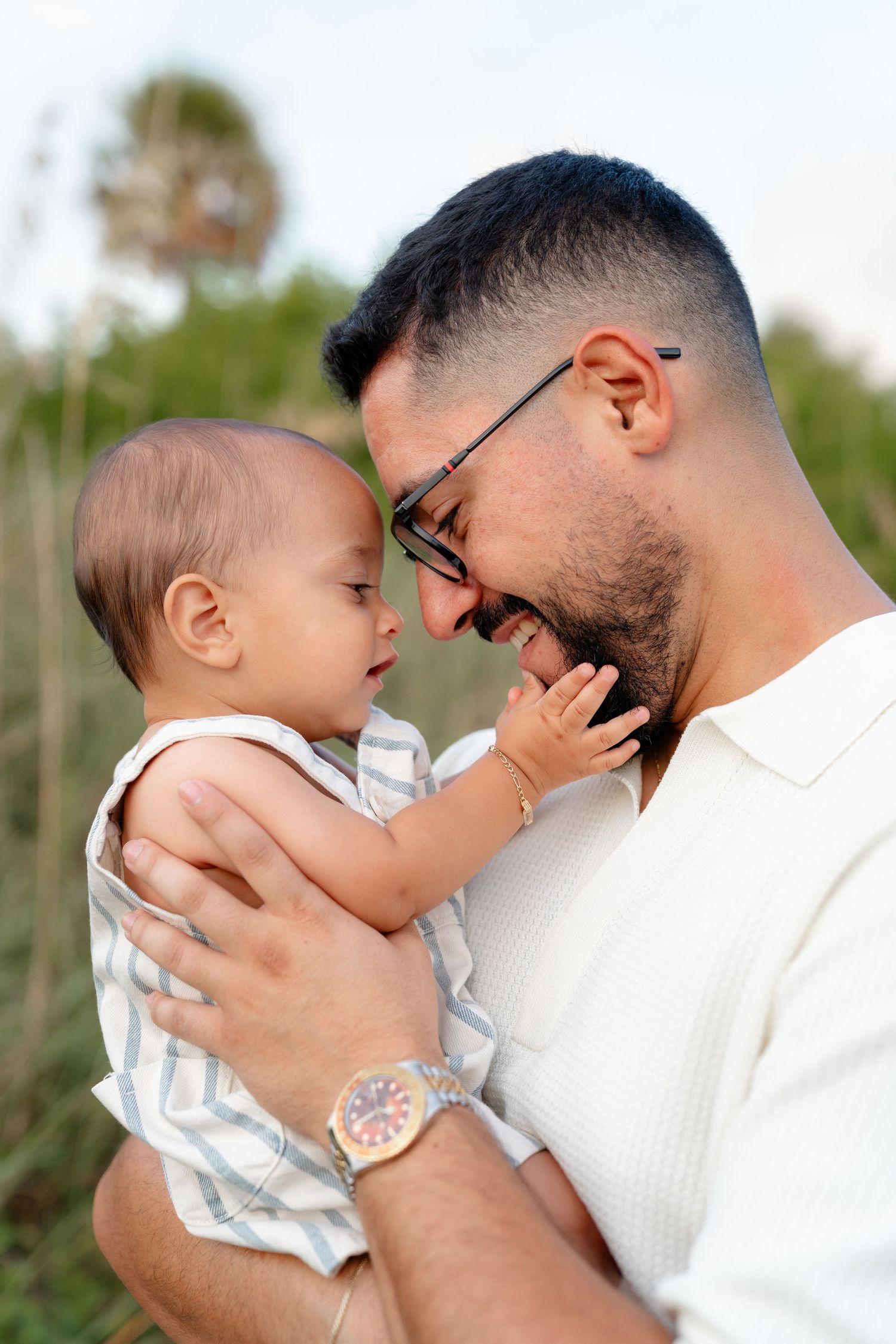 A smiling bearded father wearing glasses holds his baby outdoors, sharing a joyful face-to-face moment.