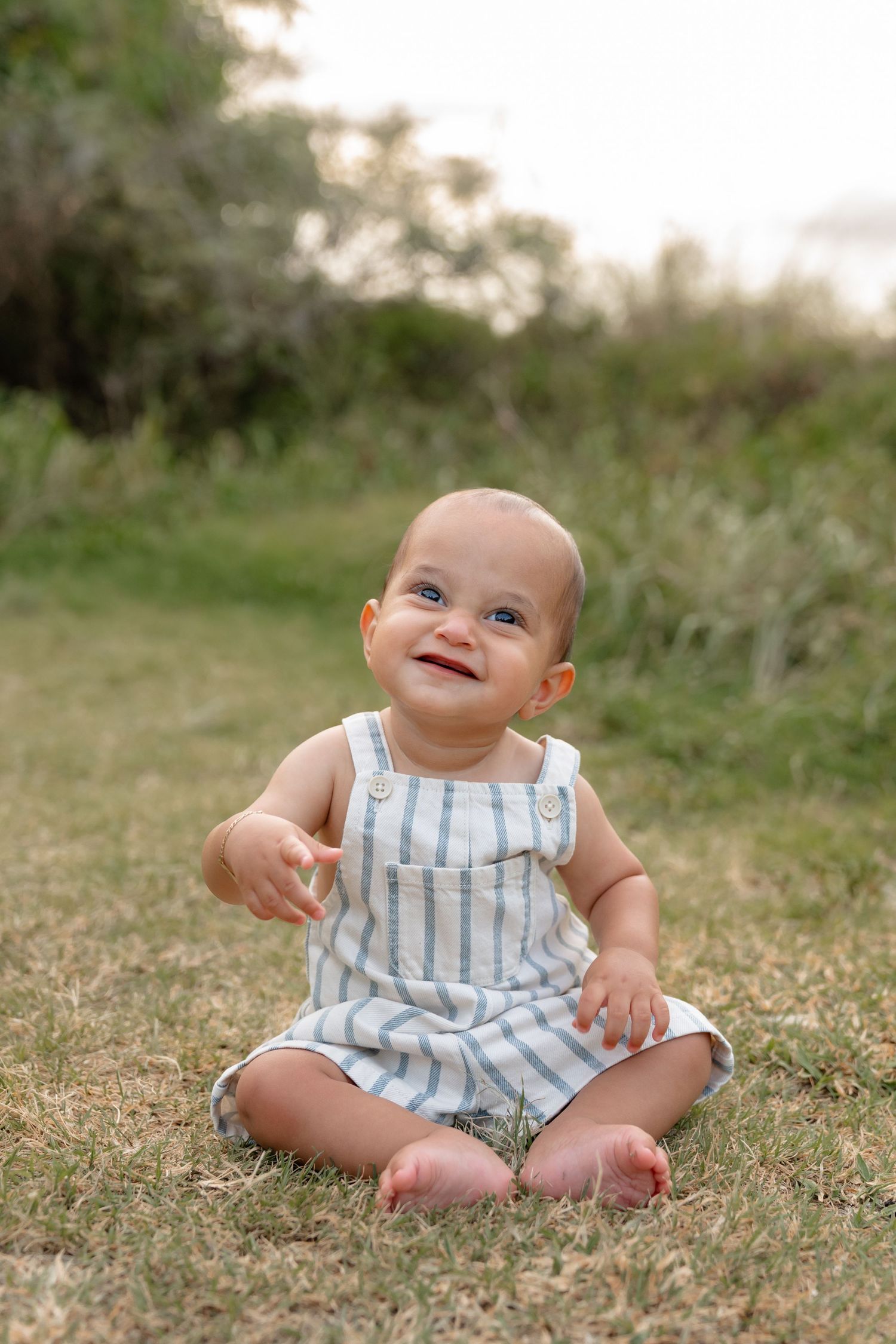 Smiling baby in striped romper sitting on green grass outdoors during golden hour.