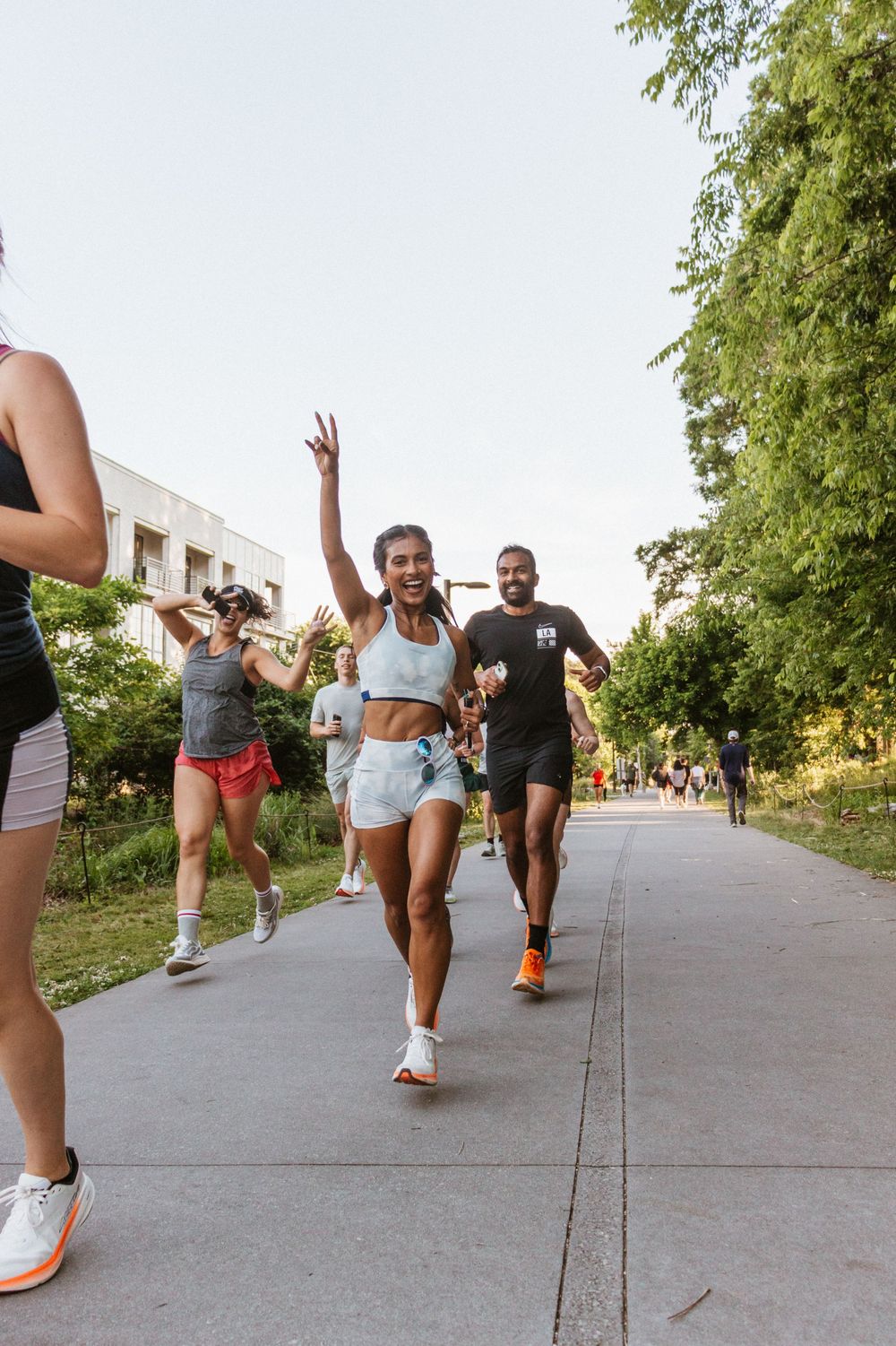 Group of diverse runners jogging on a sunny park path, with a smiling woman giving a peace sign in the foreground.