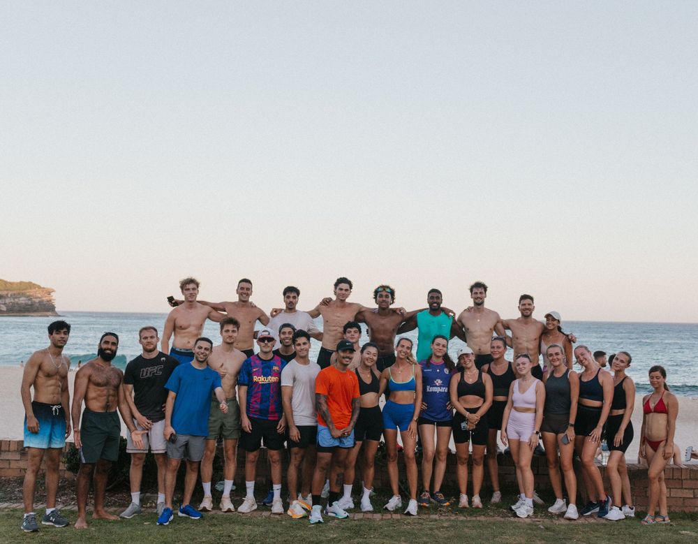 Large group of fit athletes posing together near ocean cliffs at sunset during outdoor fitness event.