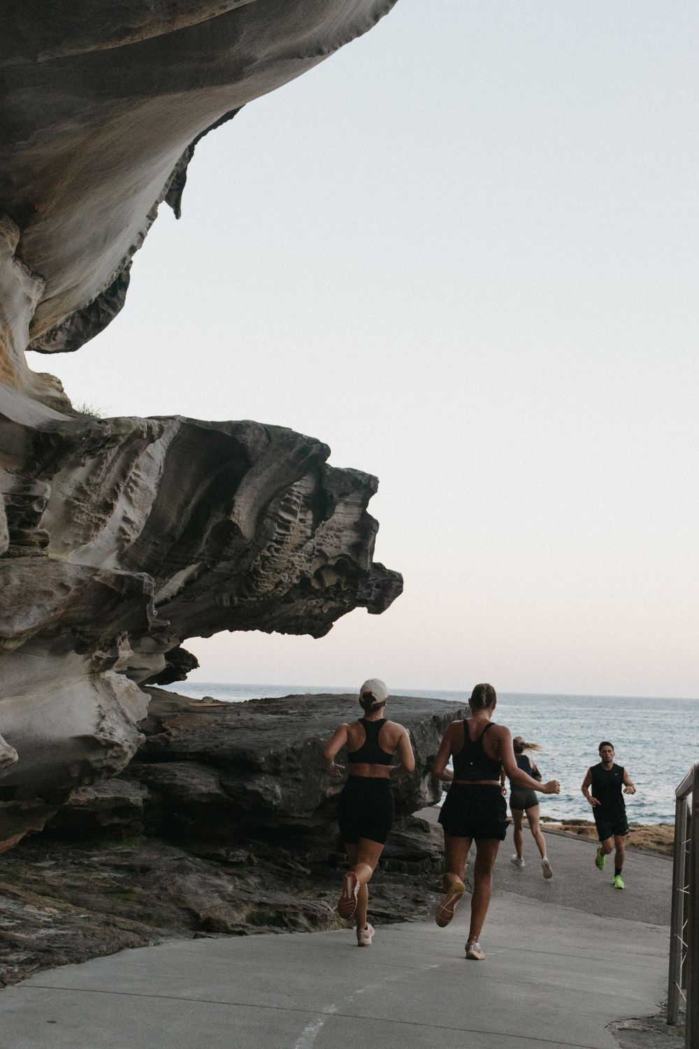 Runners in black athletic wear jogging along a coastal path beside dramatic rock formations with ocean views at dusk.