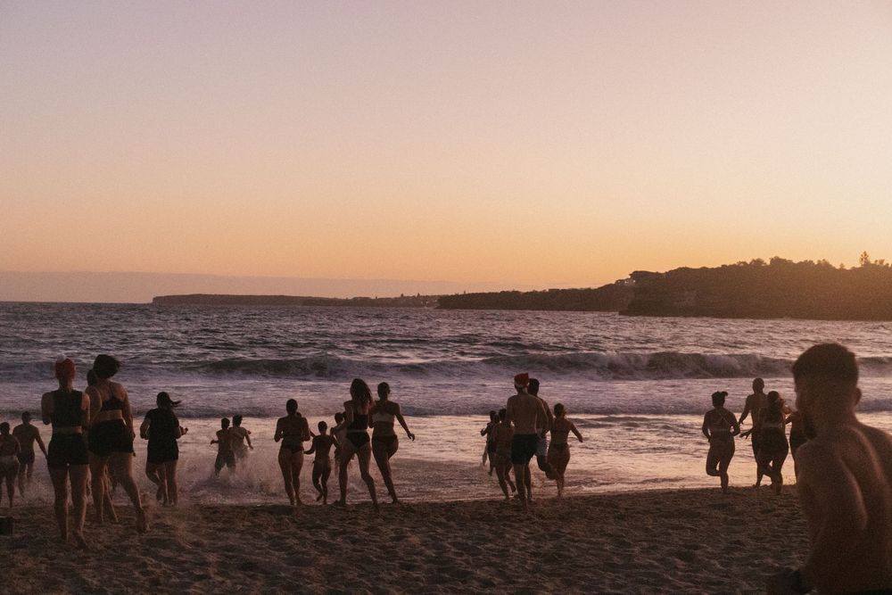 Silhouettes of people running and playing on a sandy beach at sunset with golden pink skies and ocean waves.