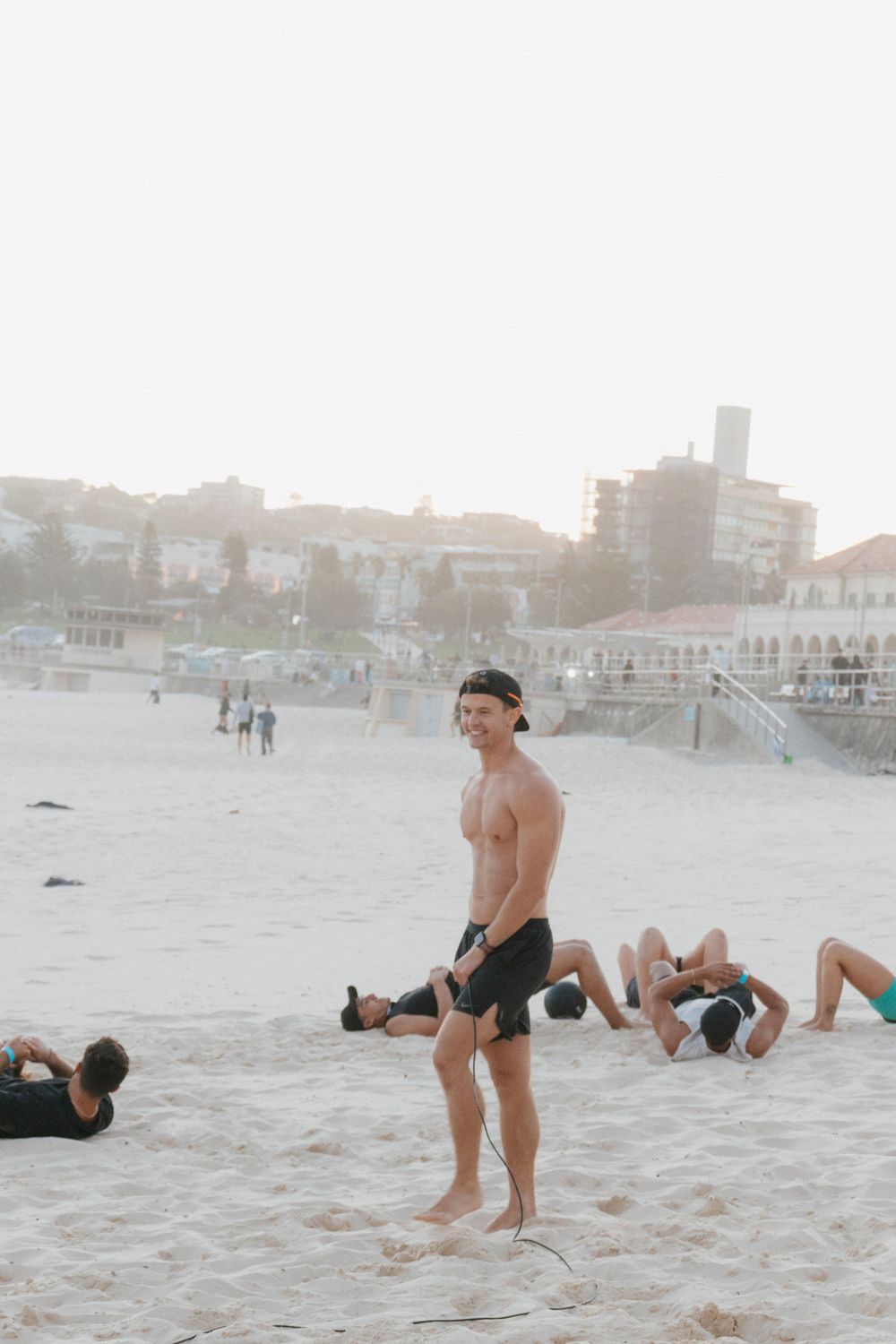Shirtless man smiling on Bondi Beach during a group fitness workout session at sunrise.