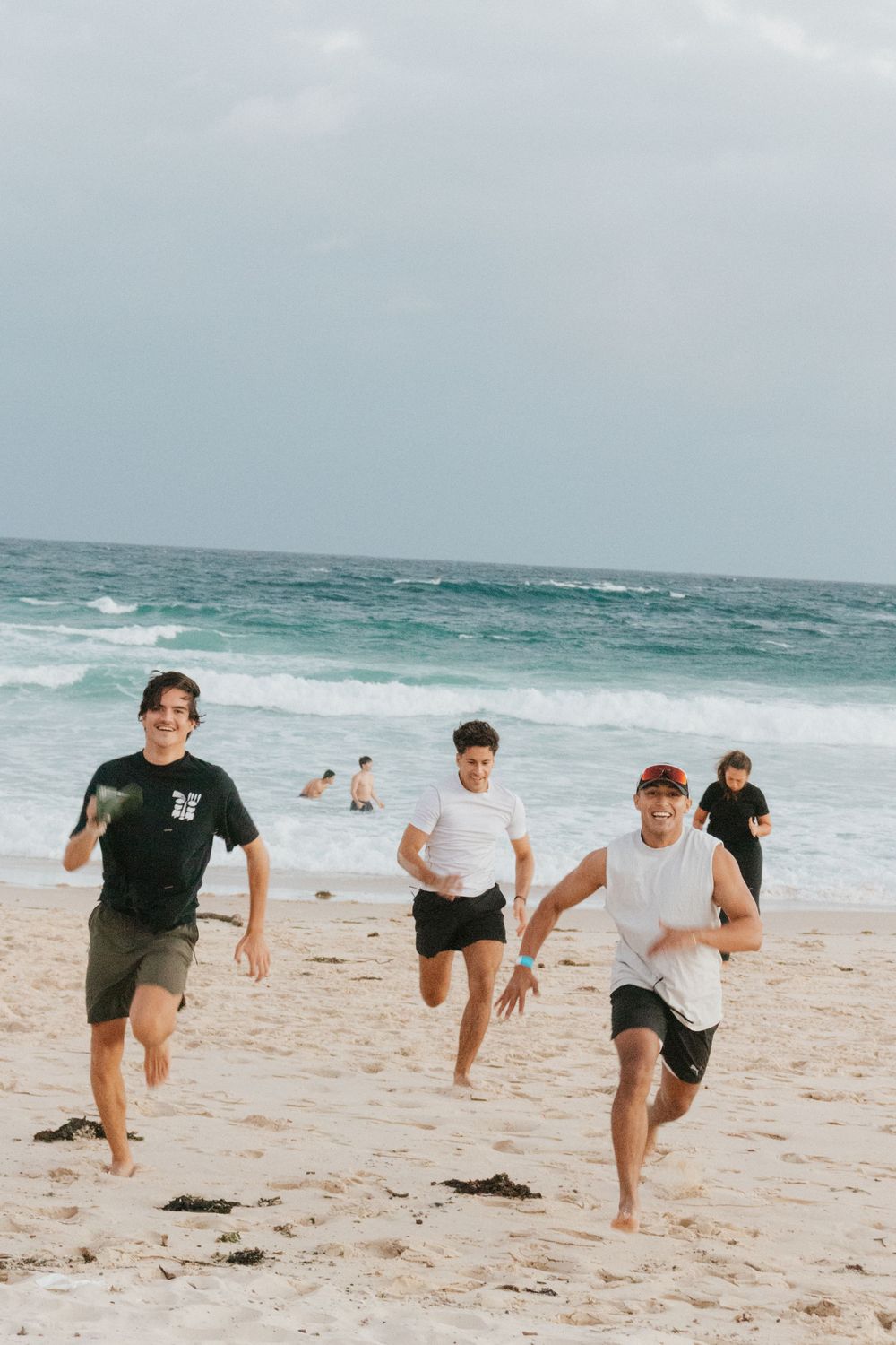 Young men running barefoot on a sandy beach with ocean waves in the background on a cloudy day.