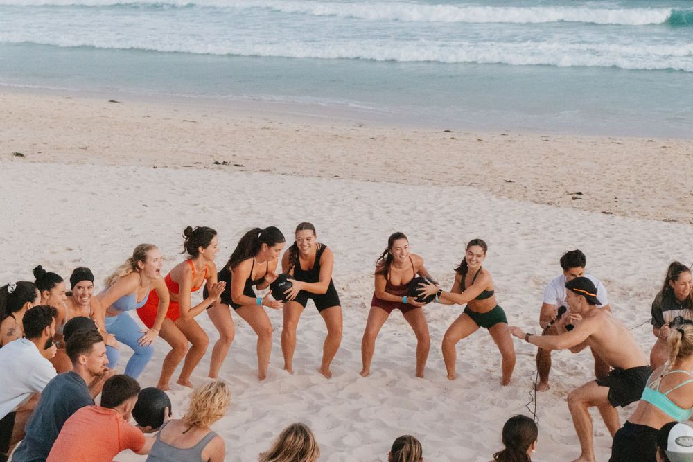 Group of women playing tug of war on a sandy beach with ocean waves in the background.
