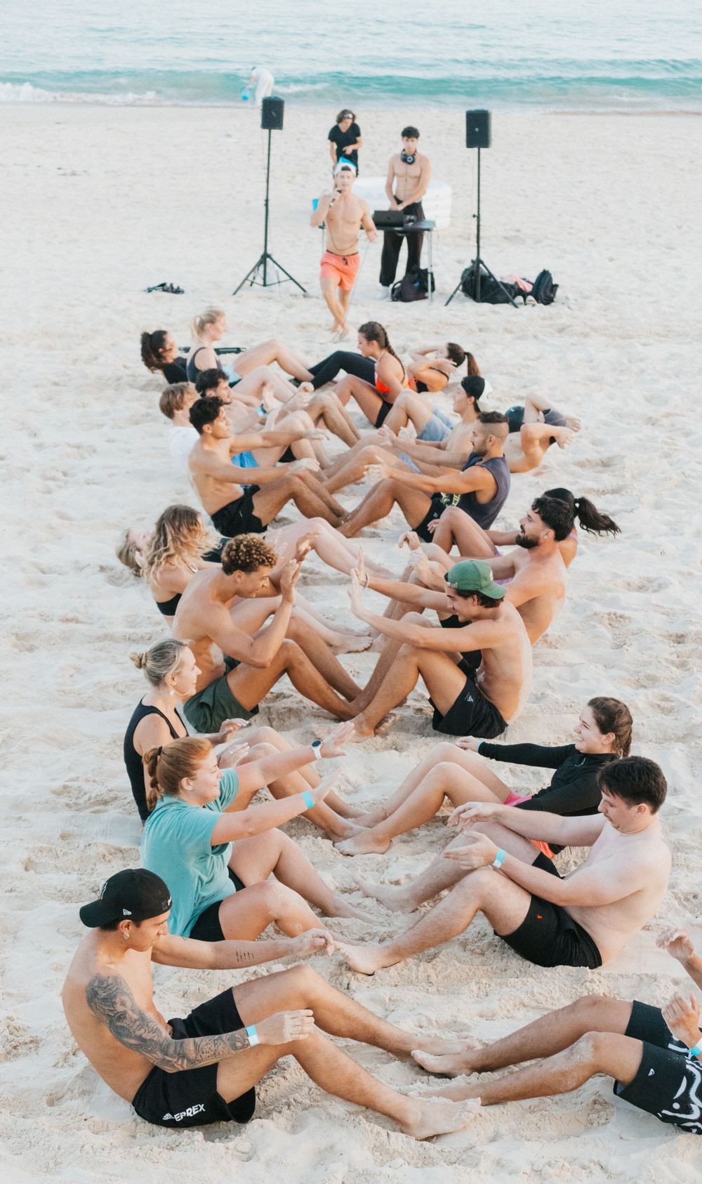 Large group doing partner sit-ups on sandy beach during outdoor fitness class with speakers and instructor in background.