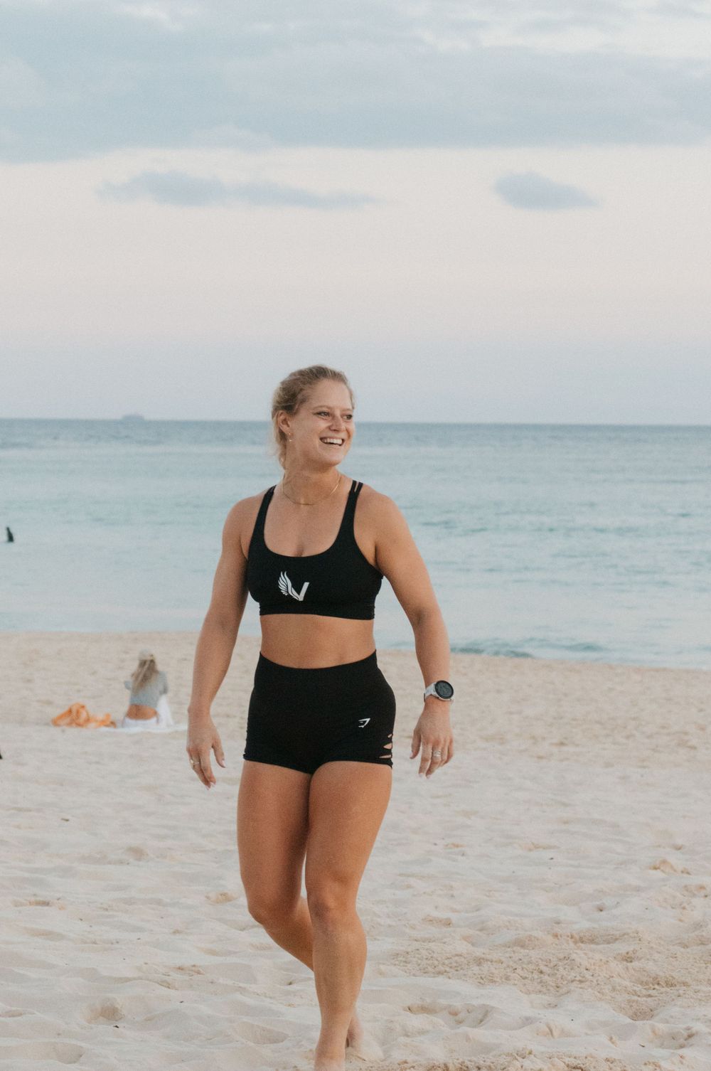 Athletic woman in black sports outfit smiling on sandy beach with ocean backdrop at dusk.
