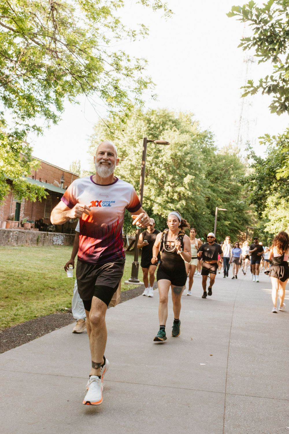 Smiling older man in race shirt leads group of runners on sunny park path during outdoor fun run event.