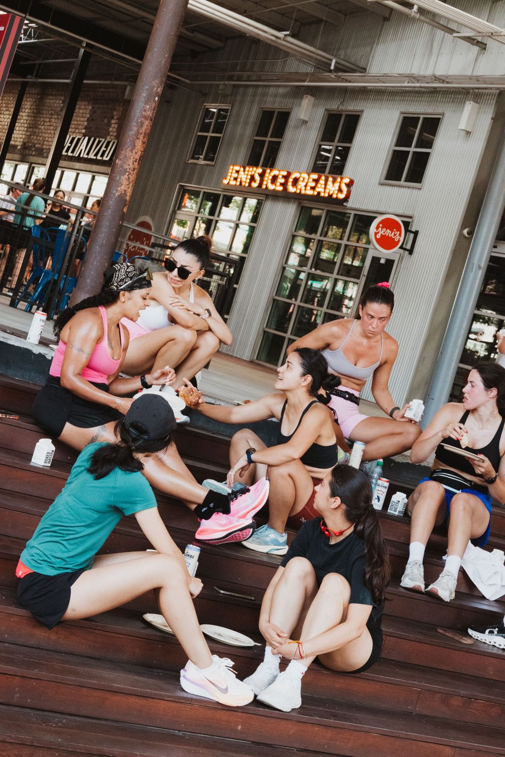 Group of women in athletic wear resting on outdoor steps near Jeni's Ice Creams shop after a workout.