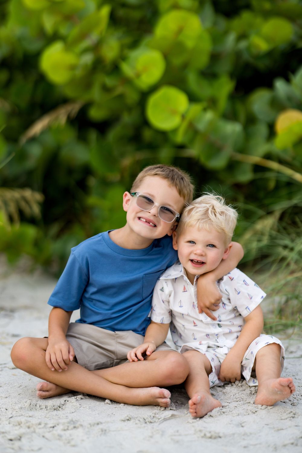 Two young brothers sitting together on sandy beach, smiling, with lush green foliage behind them.