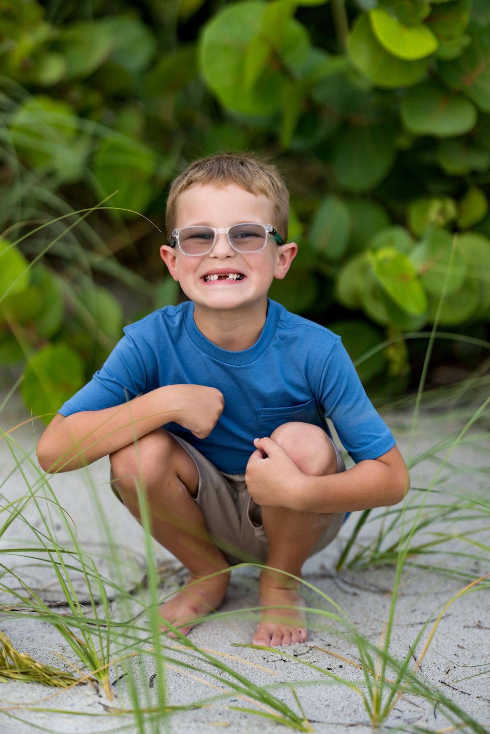 A smiling boy with glasses and missing teeth crouches on sandy beach grass wearing a blue shirt.