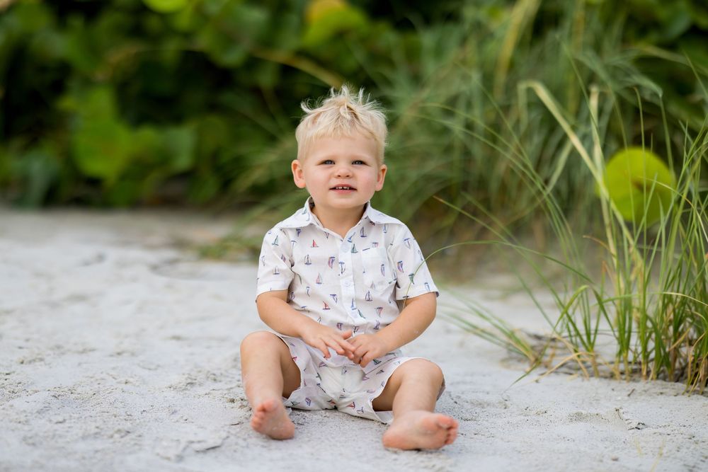 Smiling toddler boy with blonde hair sitting barefoot on sandy beach in white printed romper with tropical greenery.
