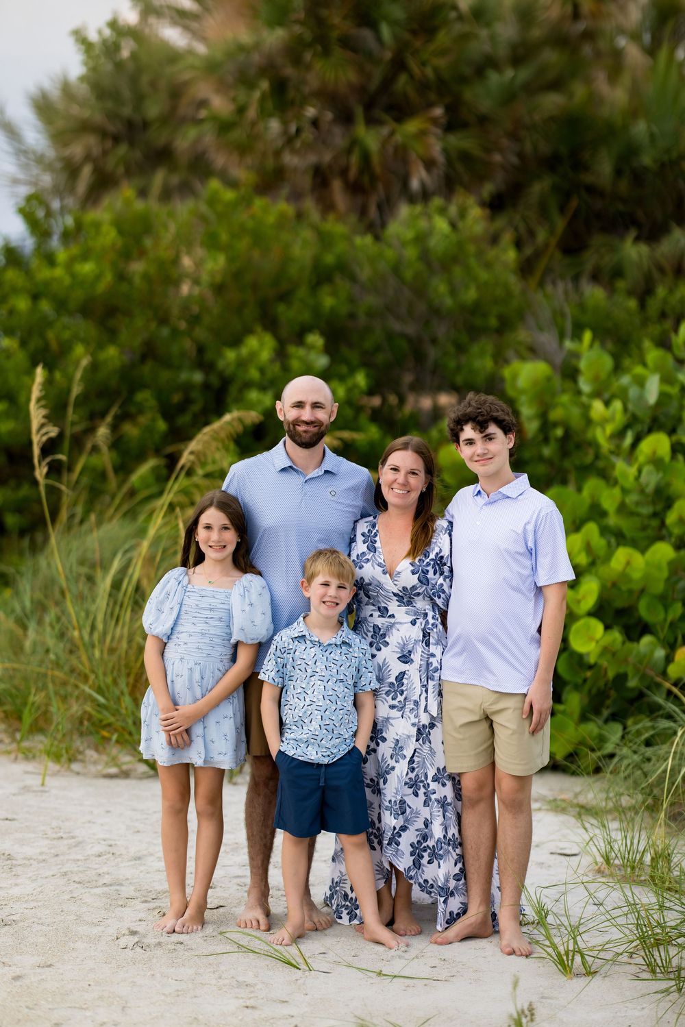 Family of five posing together on a sandy beach, dressed in coordinating blue outfits, with lush green dunes in the background.
