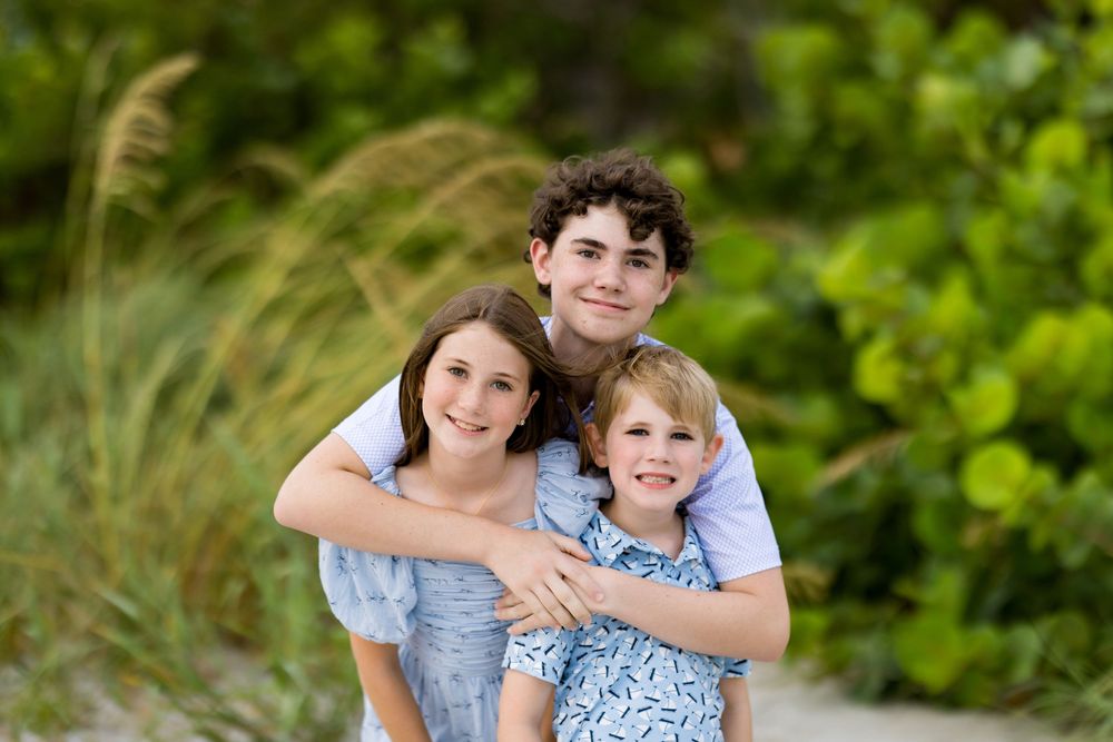 Three smiling siblings pose together on a sandy beach path surrounded by lush green tropical foliage.