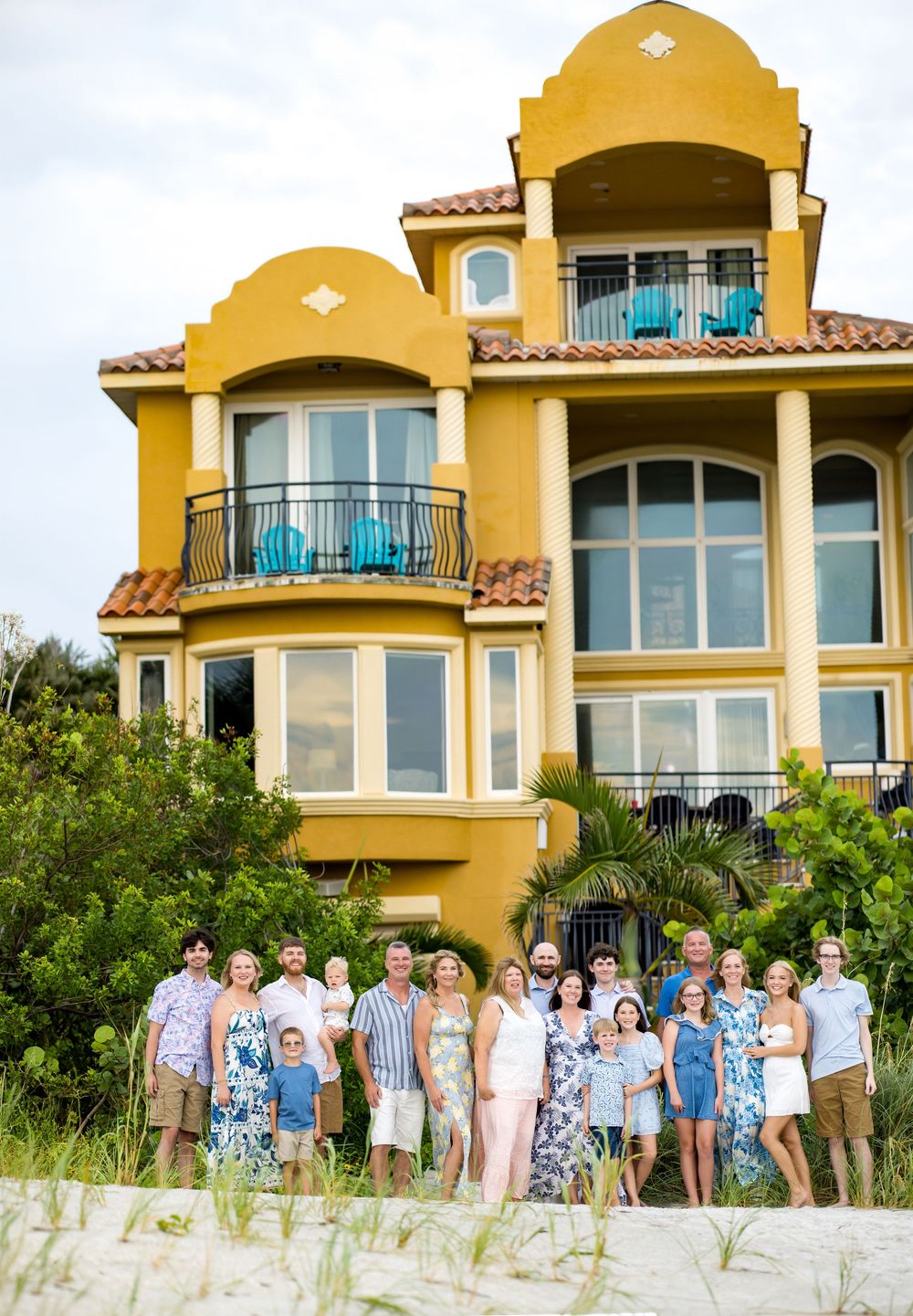 Large family group photo on beach in front of a stunning yellow Mediterranean-style three-story vacation home.