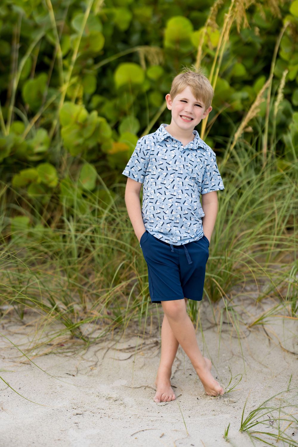 Smiling blonde boy in blue patterned shirt and navy shorts standing barefoot on sandy beach dunes with green sea oats.