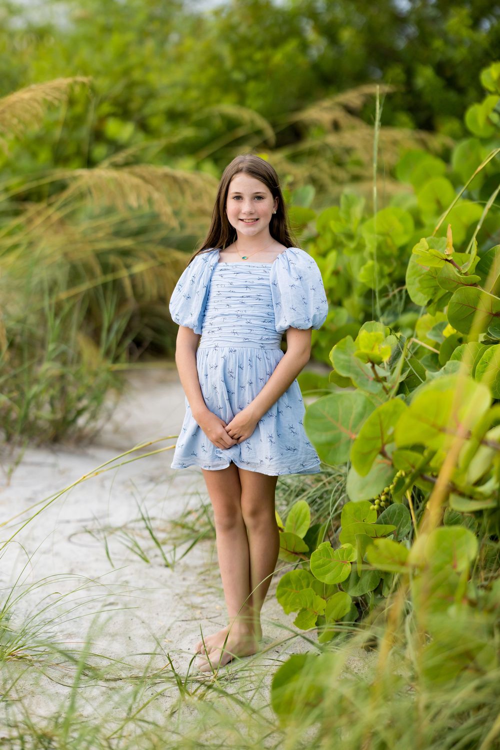Young girl in blue puff-sleeve dress standing on sandy beach path surrounded by lush green tropical foliage.
