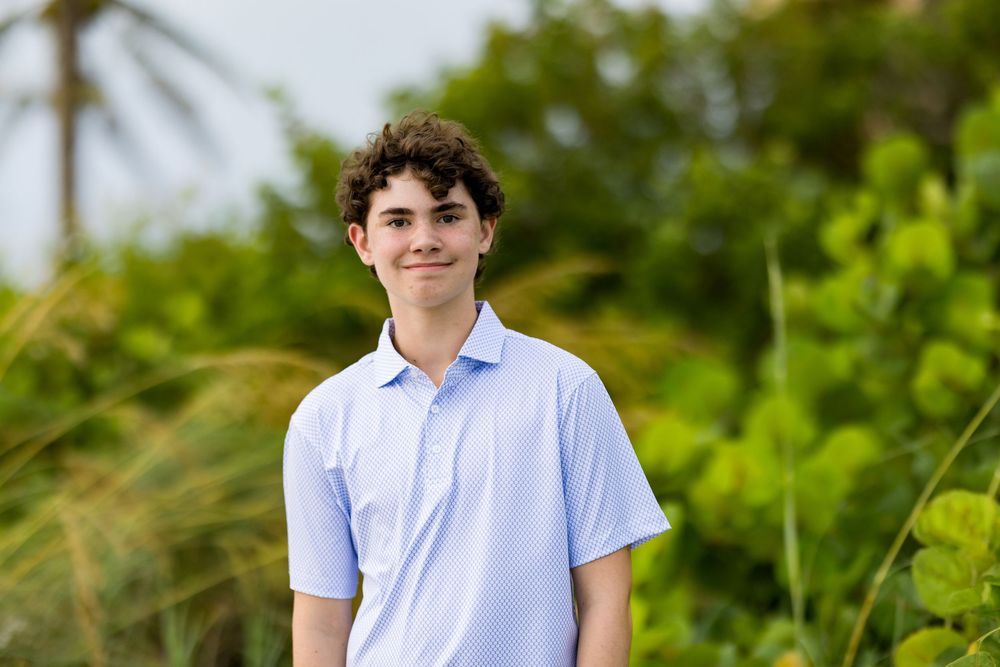 Smiling teenage boy with curly hair wearing a light blue polo shirt outdoors with lush green tropical foliage.