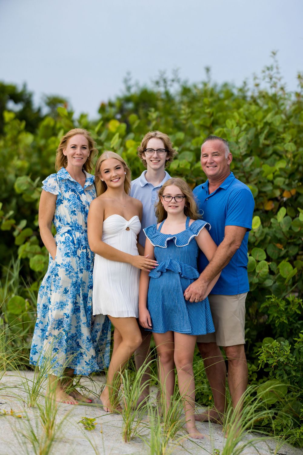 Family of five posing together on sandy beach dunes with lush green foliage, wearing coordinated blue and white outfits.