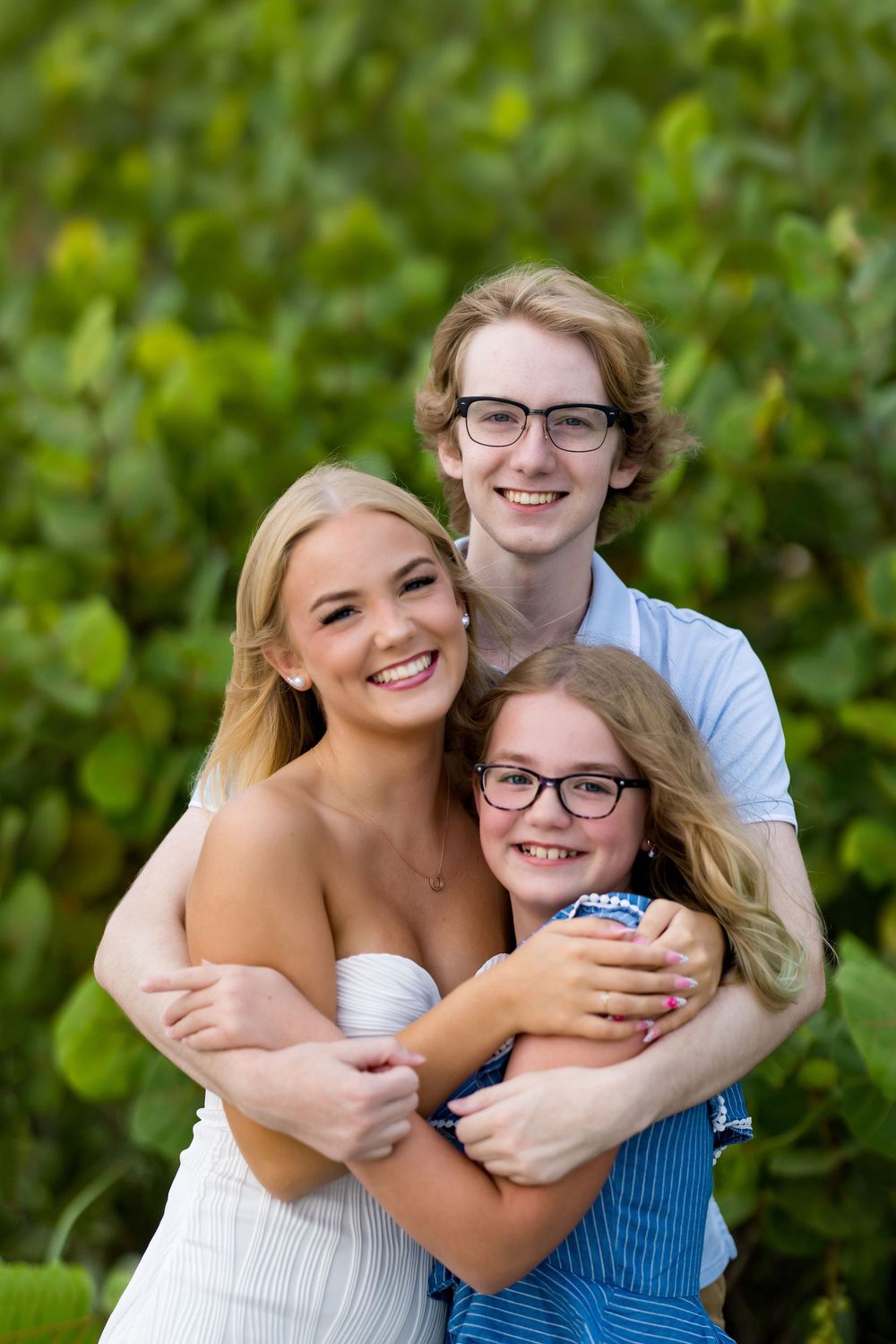 Three smiling siblings hugging outdoors with lush green foliage background, two teens and a young girl in casual summer attire.