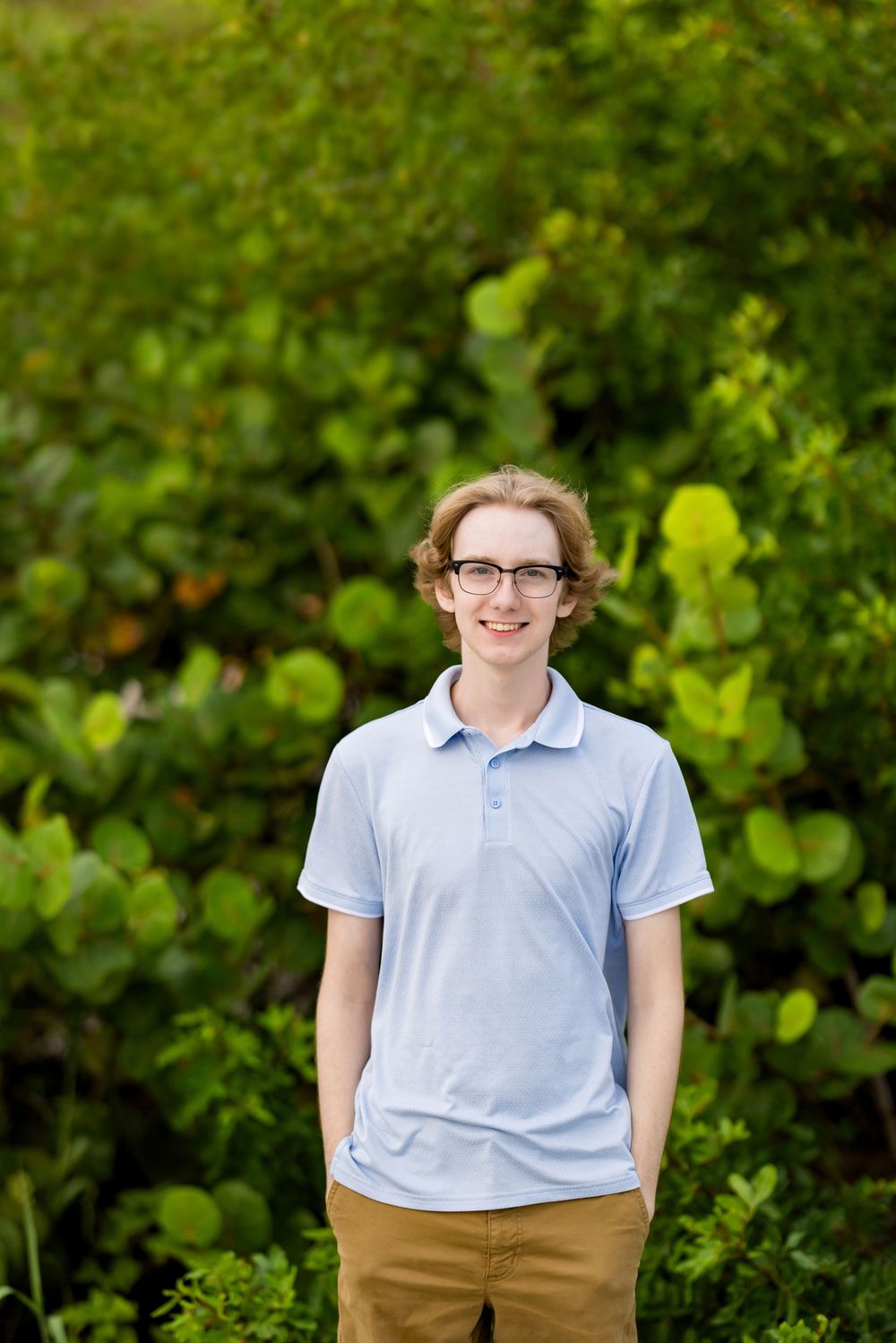 Smiling young man with glasses in light blue polo shirt standing outdoors in front of lush green foliage.