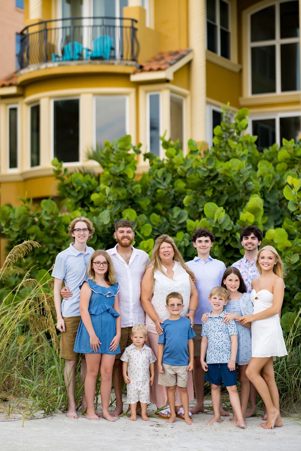 Large family group portrait on sandy beach in front of colorful vacation home with lush tropical greenery.
