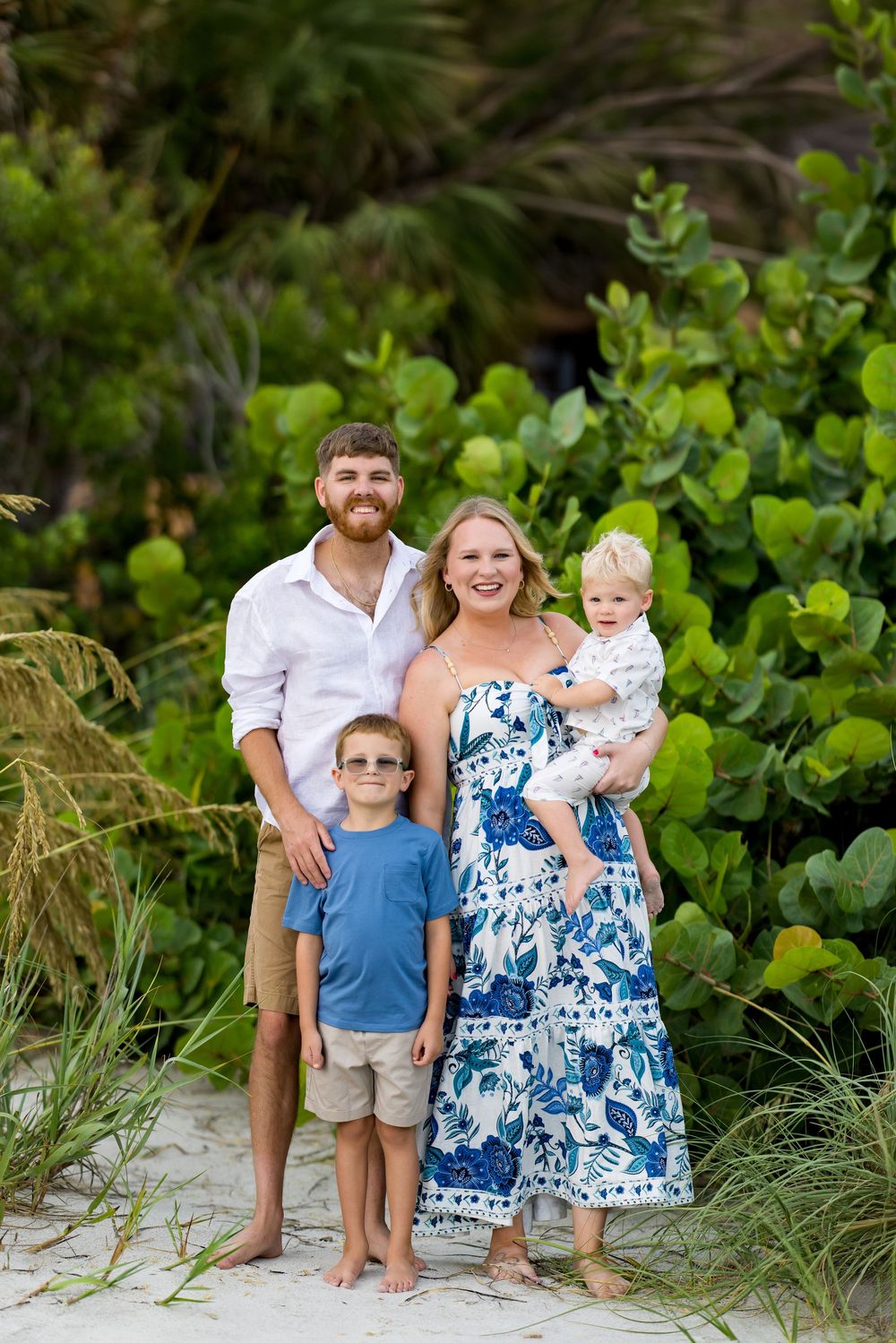 Family of four poses on sandy beach path surrounded by lush green tropical foliage for a portrait photo.