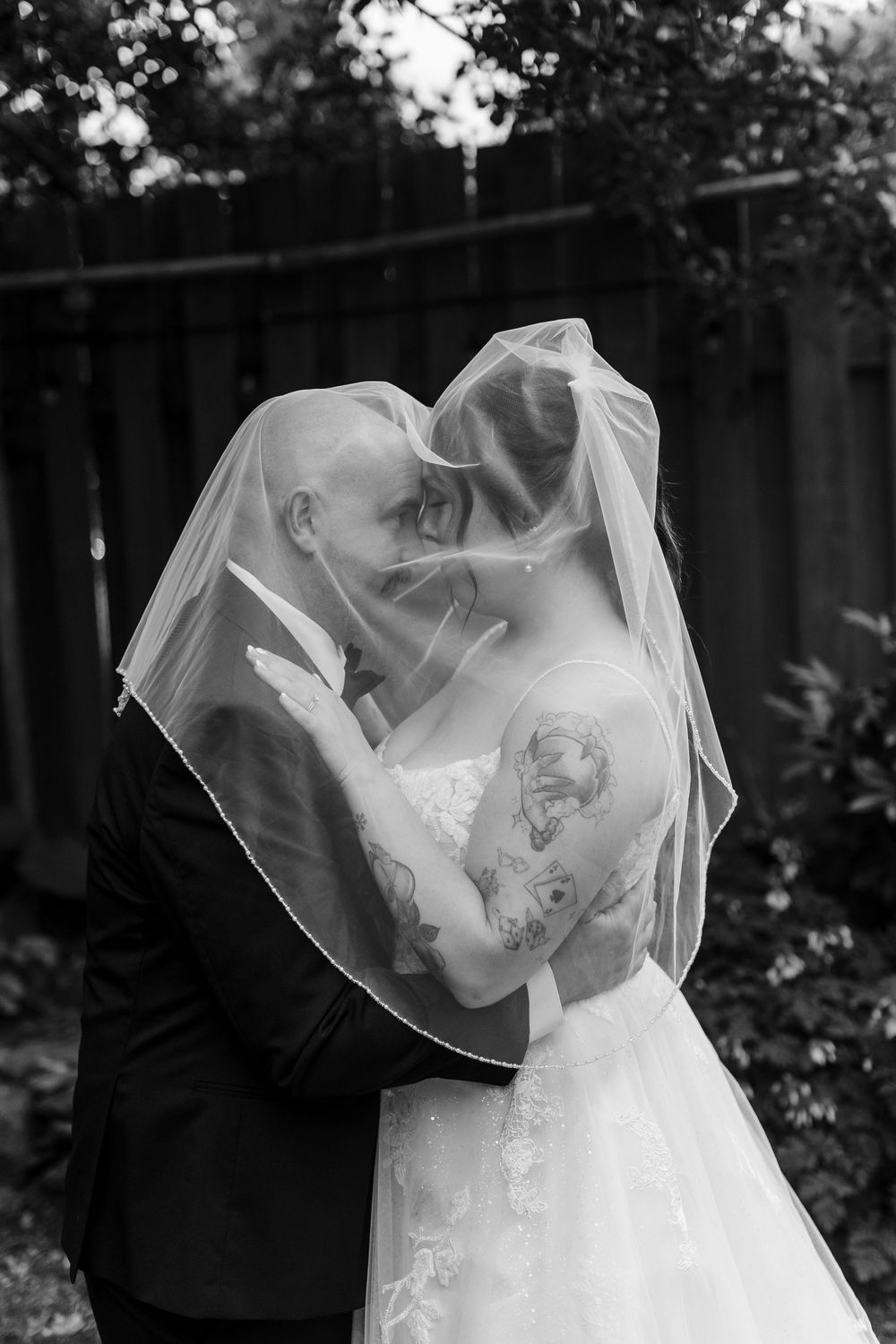 Black and white photo of bride and groom kissing under wedding veil outdoors, bride has tattoos on her arm.